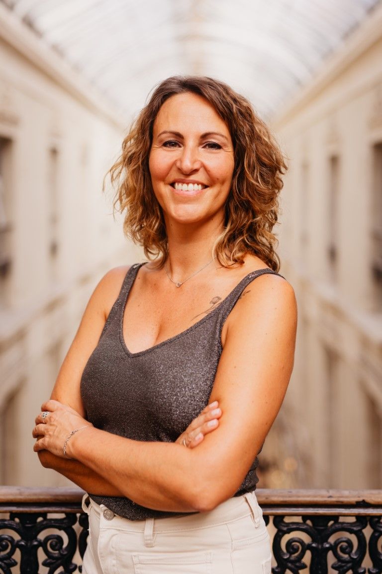 Smiling woman with curly brown hair standing with arms crossed in elegant arcade.