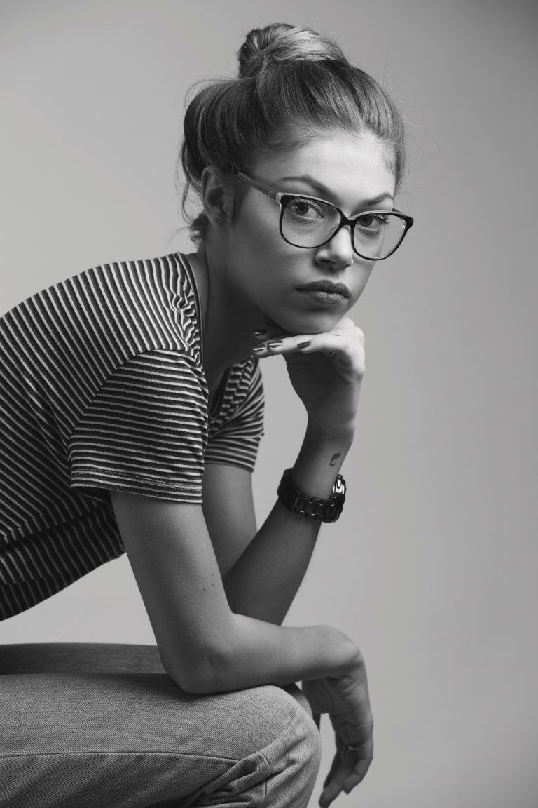 Photo en noir et blanc d'une jeune femme portant des lunettes, un chignon et un haut rayé, posant d'un air pensif.