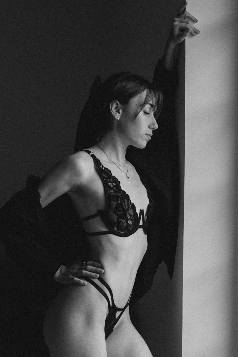 Black and white photo of a slender woman in black lingerie leaning on a wall.