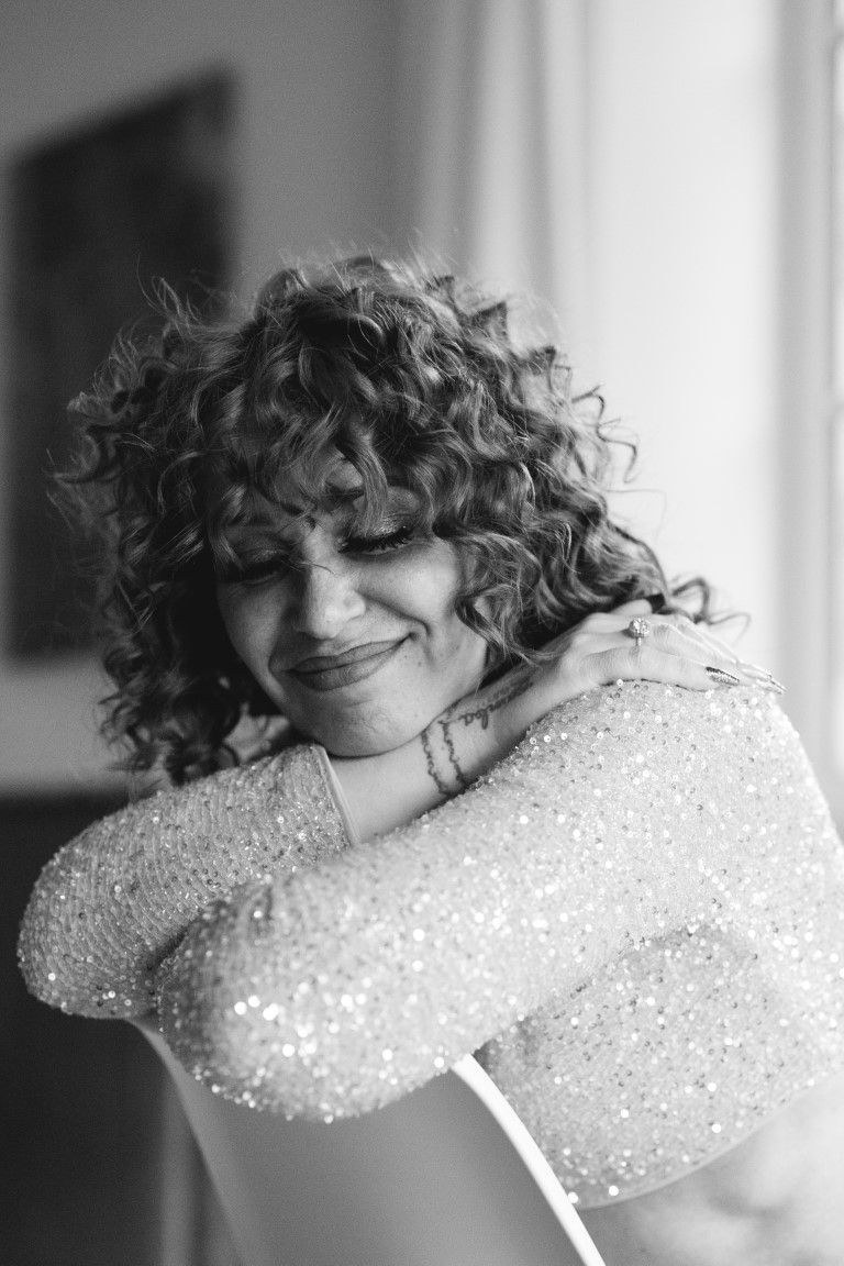 Black and white portrait of curly-haired woman smiling with arms crossed in sparkly top.