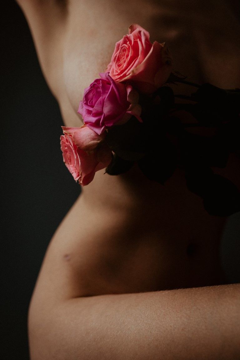 Gros plan sur des roses roses et corail posées contre le torse nu d'une femme, dans une lumière sombre et dramatique.