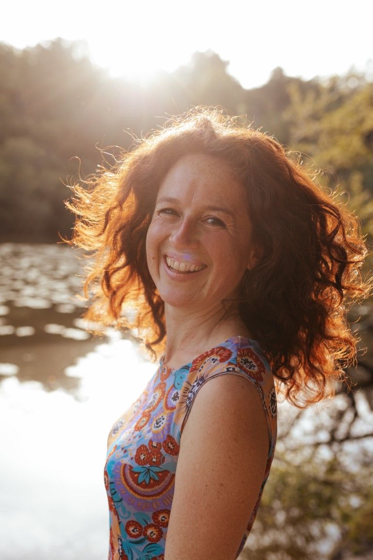 Smiling woman with curly red hair wearing a floral dress, standing outdoors near a sunlit lake.