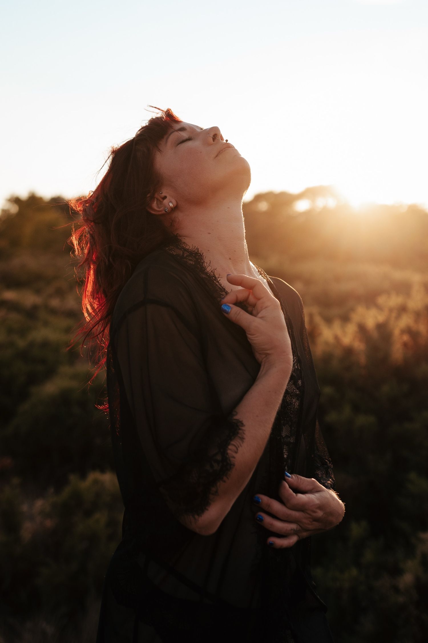 Woman in black dress tilting head back at golden sunset in a field, hair glowing with warm backlight.