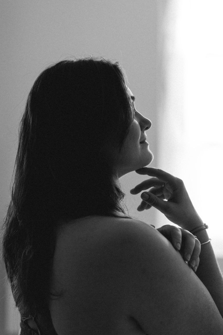Black and white silhouette profile of woman with long hair touching chin near bright window.