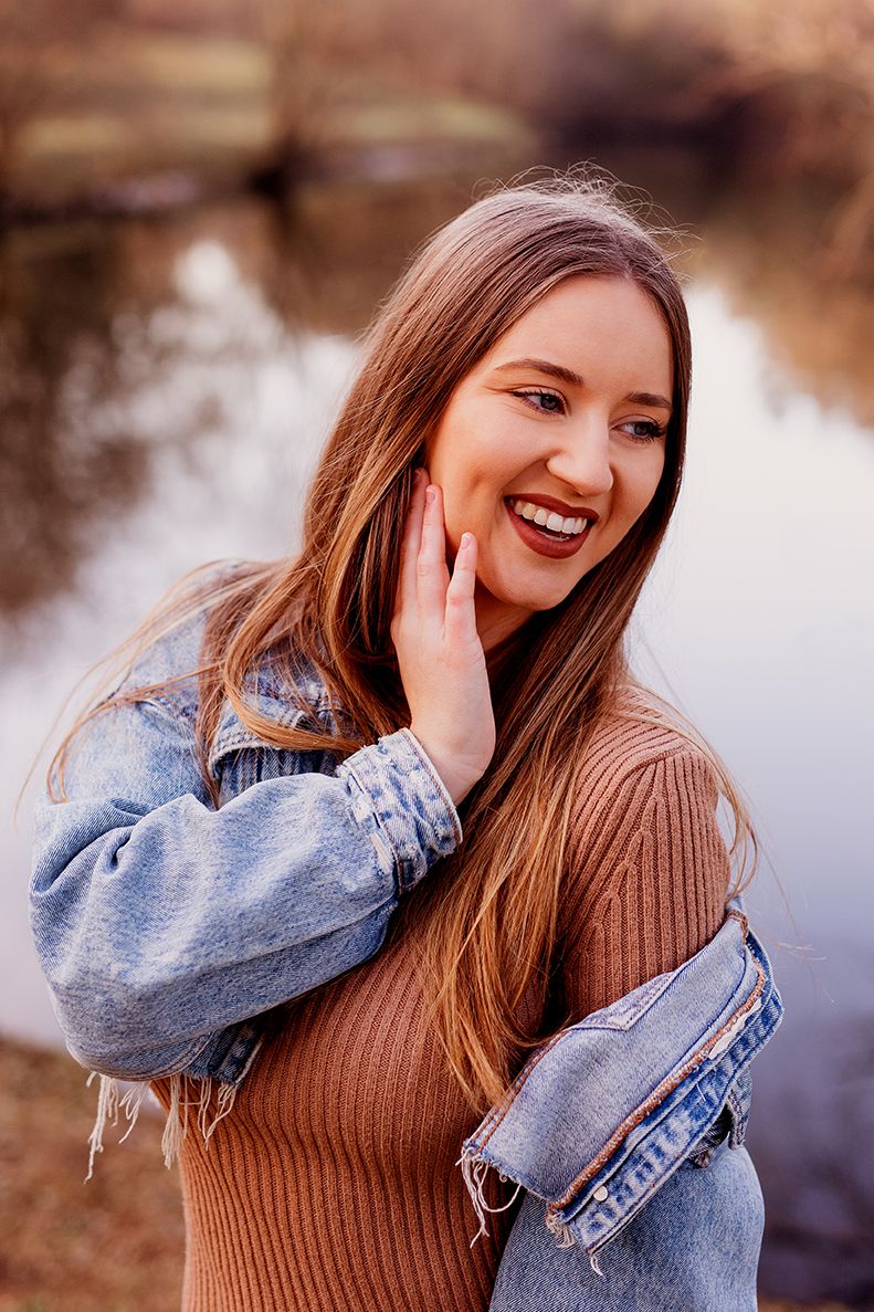 Jeune femme souriante en veste en jean et pull marron, riant au bord d'un étang réfléchissant à Granville