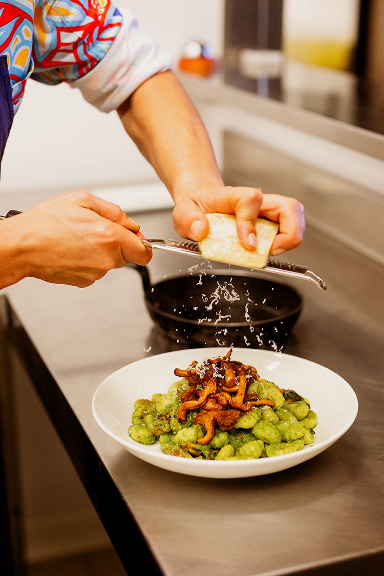 Chef grating parmesan cheese over green gnocchi with chanterelle mushrooms in white bowl.