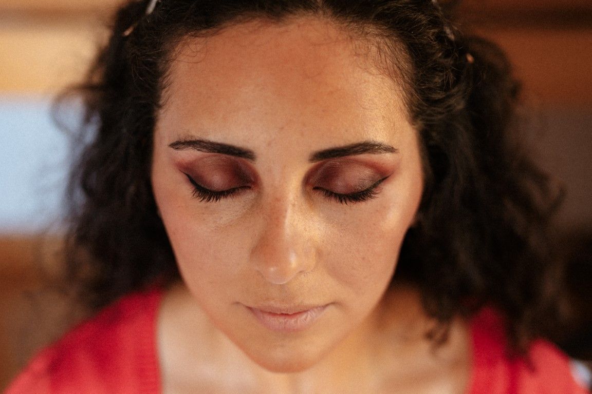 Close-up portrait of a woman with curly dark hair and elegant eye makeup, eyes closed, wearing a red top.