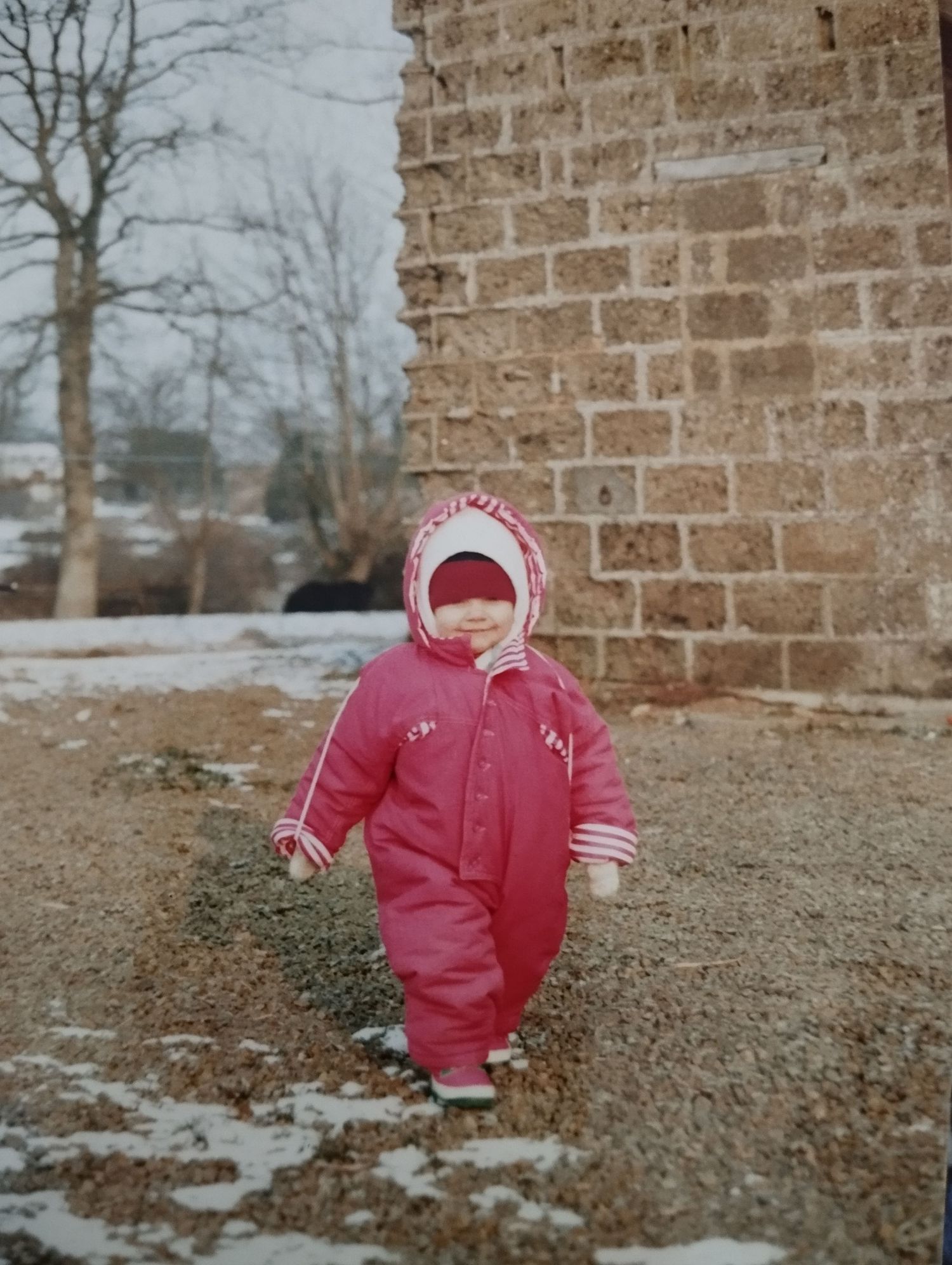 Un enfant en bas âge vêtu d'une combinaison de neige rose se tient à l'extérieur, près d'un muret de pierre, sous une légère neige hivernale