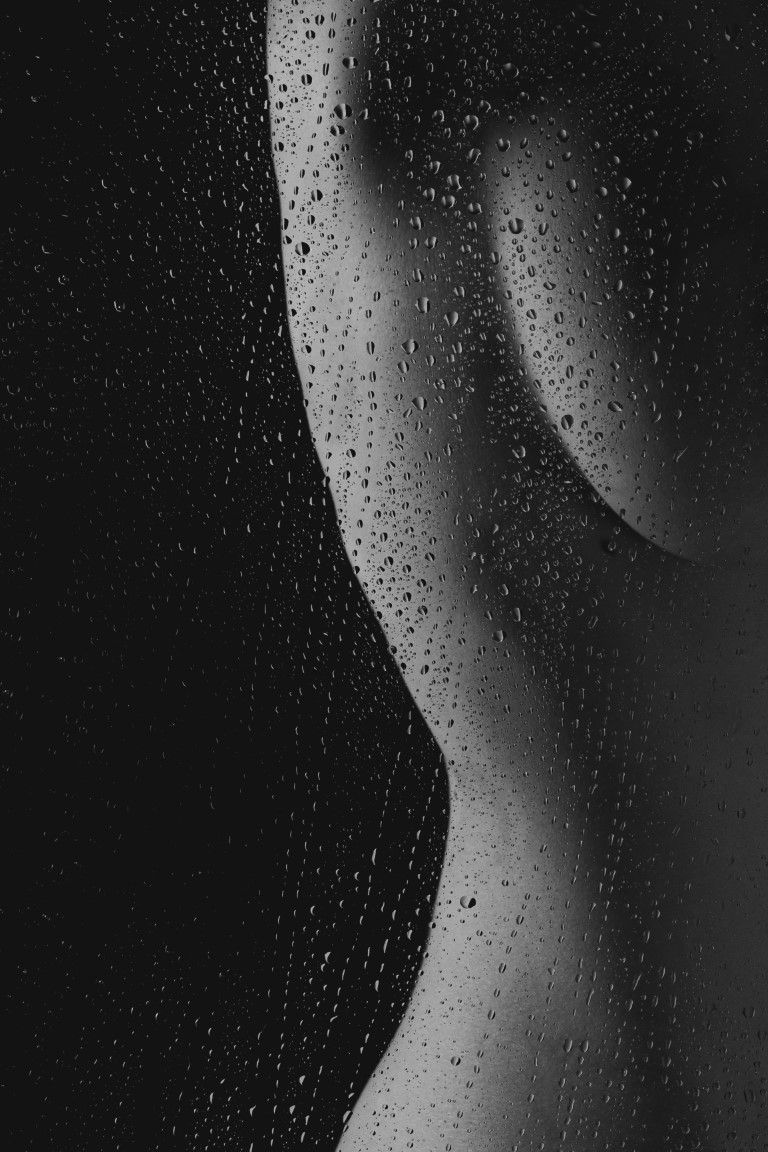 Silhouette artistique en noir et blanc d'un nu photographié à travers une vitre recouverte de pluie, sous un éclairage dramatique.