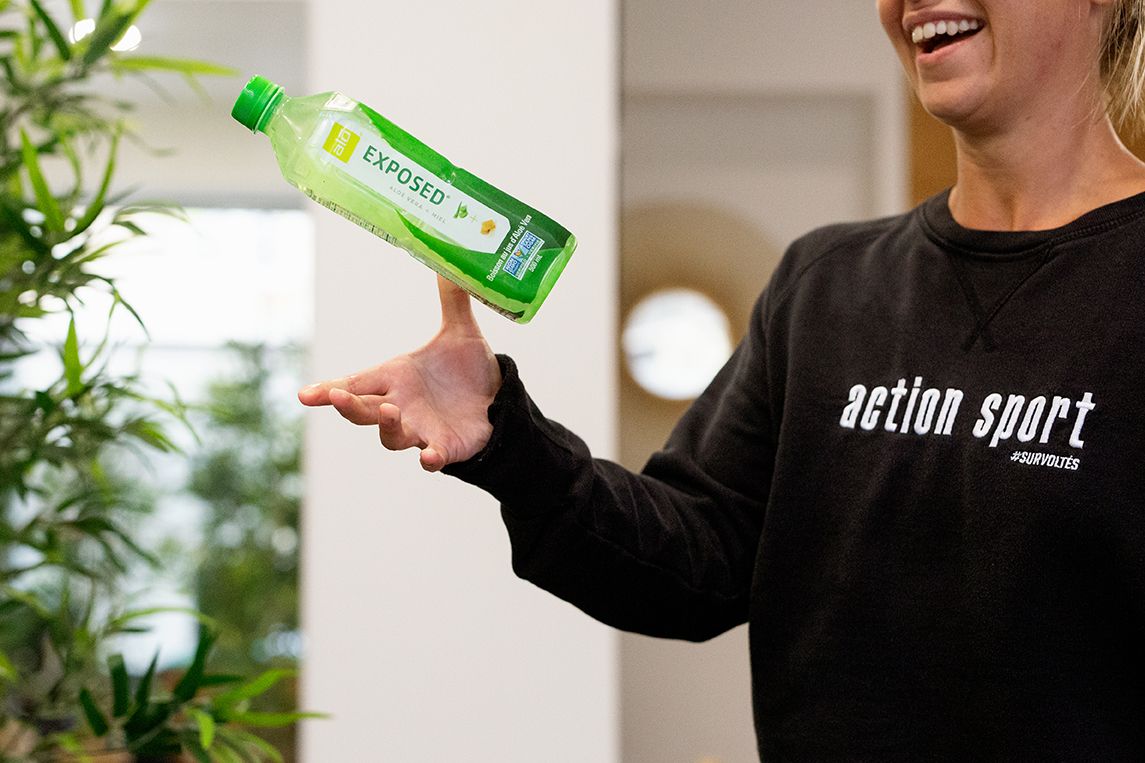 Smiling woman in black Action Sport sweatshirt balancing green Deposa bottle on finger.