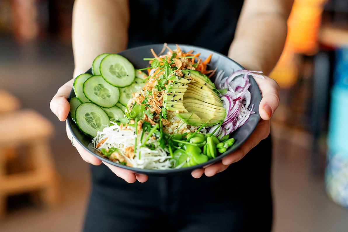 Hands holding a dark bowl filled with fresh avocado, cucumber, and mixed greens.
