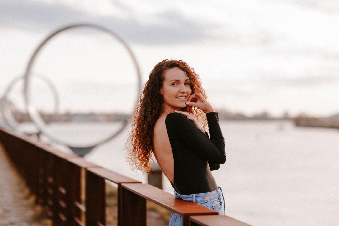 Femme aux cheveux bouclés, vêtue d'un haut noir, souriant sur un pont au bord de l'eau au coucher du soleil. à Nantes