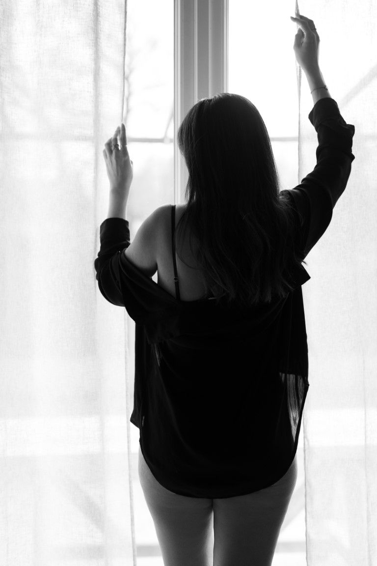 Silhouette of a woman in black lingerie standing by a sunlit window with sheer curtains in black and white.