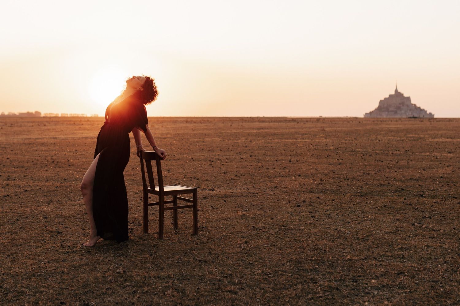 Woman in black dress leans on chair in vast field at sunset with Mont Saint-Michel silhouette in background.