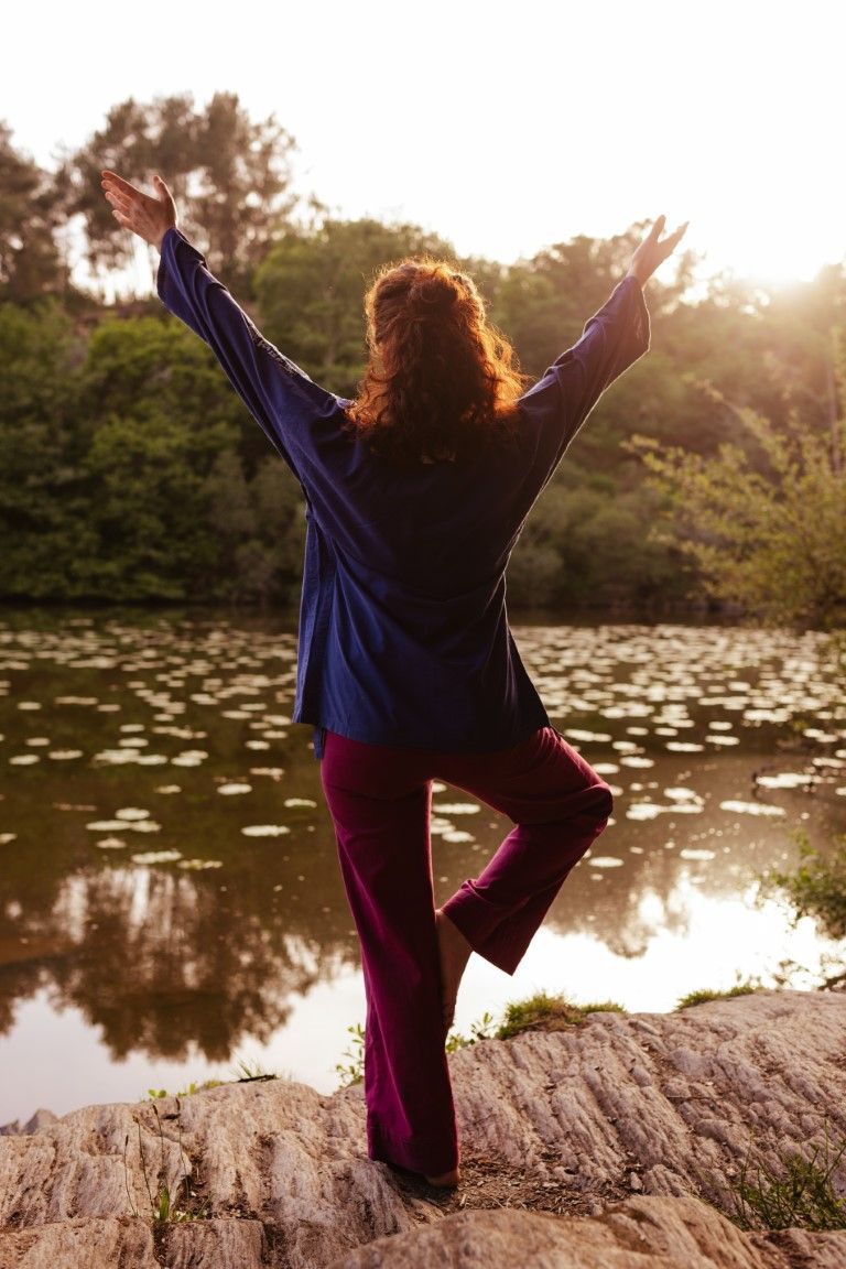 Woman in blue top and purple pants practicing tai chi by a serene lake at sunset, arms raised in graceful pose.