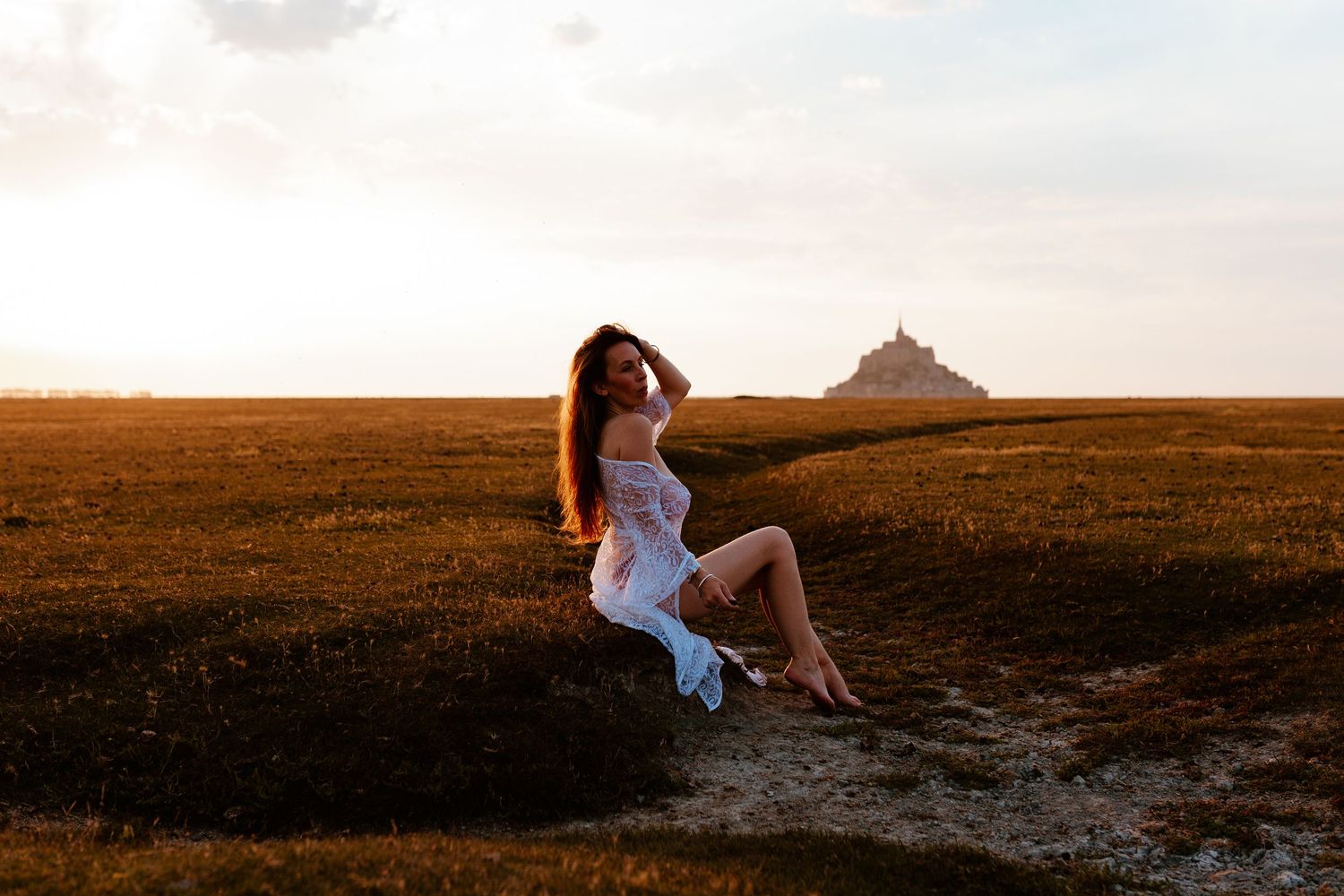 Woman in white dress sitting on rocks at sunset with Mont Saint-Michel castle in the background.