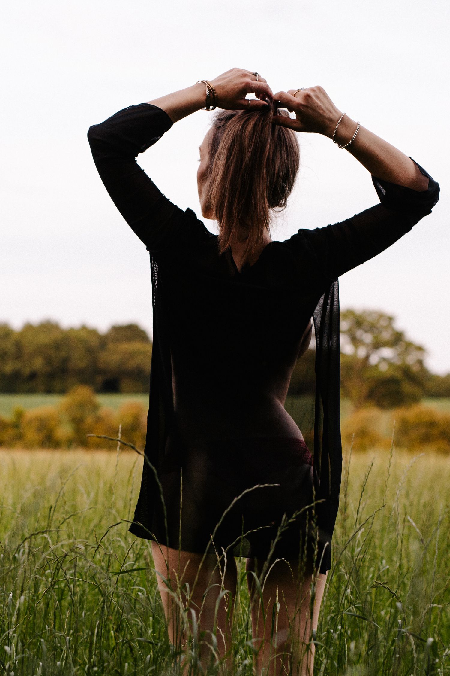 Woman in sheer black dress standing in green field, viewed from behind with hands in hair.