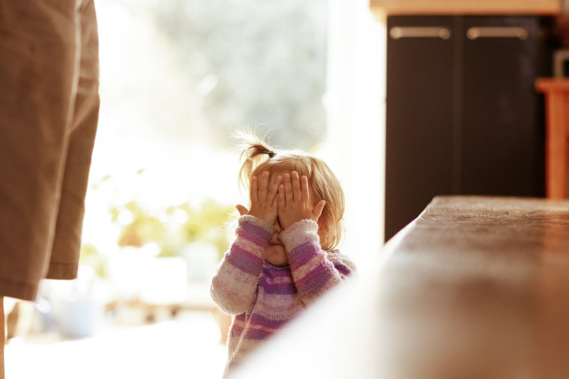 Jeune fille en pull rayé violet cachant son visage derrière ses mains, appuyée sur une table près d'une fenêtre lumineuse.