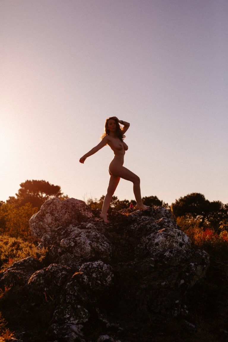 Silhouette d'une femme nue posant au sommet d'une colline rocheuse, se détachant sur un ciel doré et chaud au coucher du soleil.