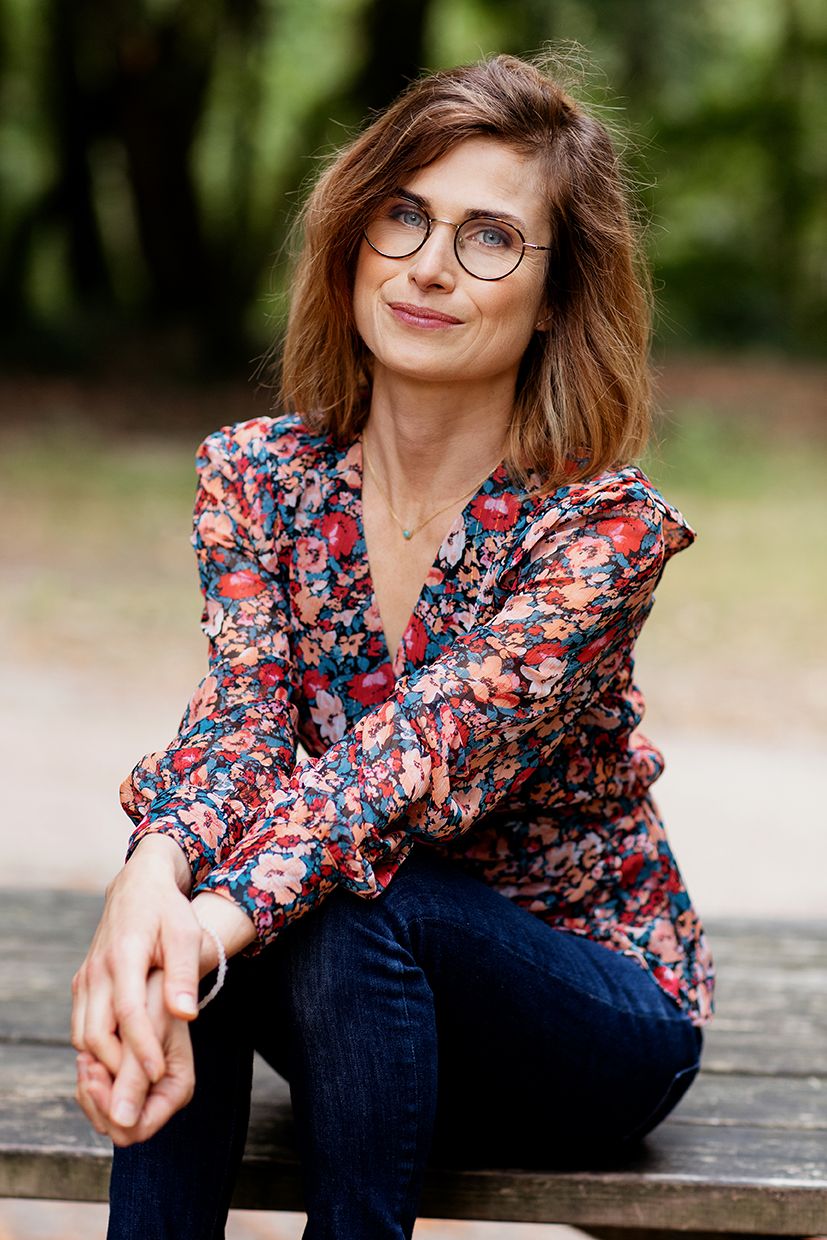 Woman in floral blouse and glasses sitting on bench outdoors, smiling in a park setting.