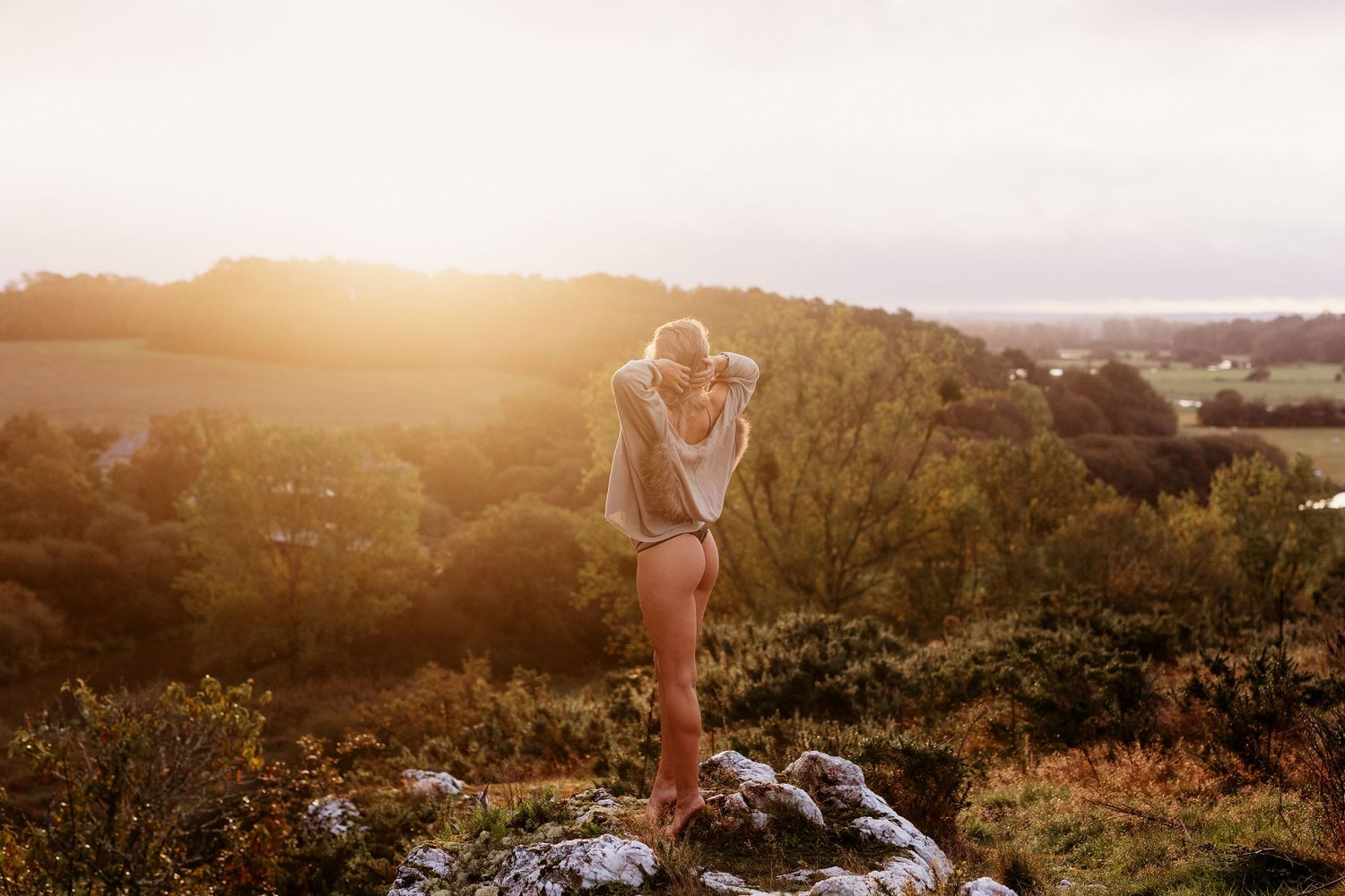 Woman standing on a rocky hilltop at golden sunset, overlooking lush green countryside landscape.