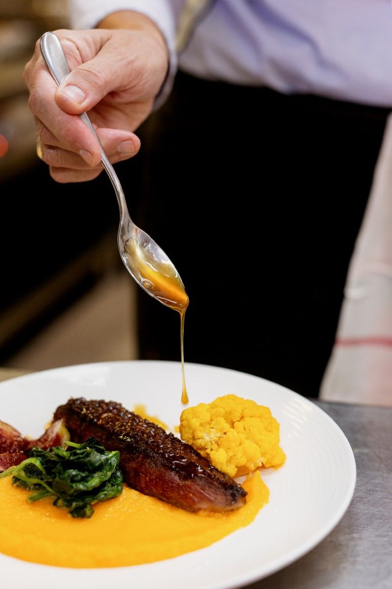 Chef drizzling golden sauce over a gourmet meat dish with vegetables on a white plate.