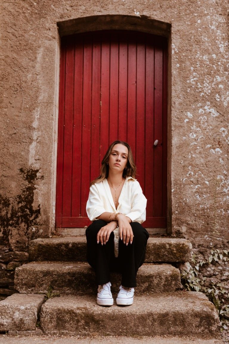 Jeune femme en chemise blanche assise sur des marches en pierre devant une porte en bois rouge.
