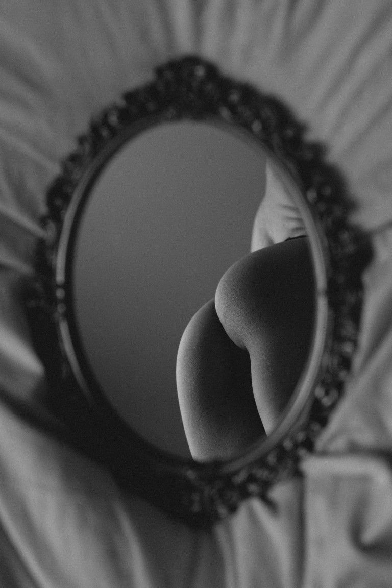 Reflet en noir et blanc des courbes nues d'une femme dans un miroir ovale orné, posé sur une literie blanche.