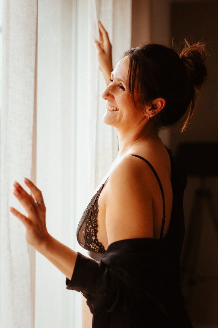 Smiling brunette woman in black lace bralette and robe standing by a bright sunlit window.