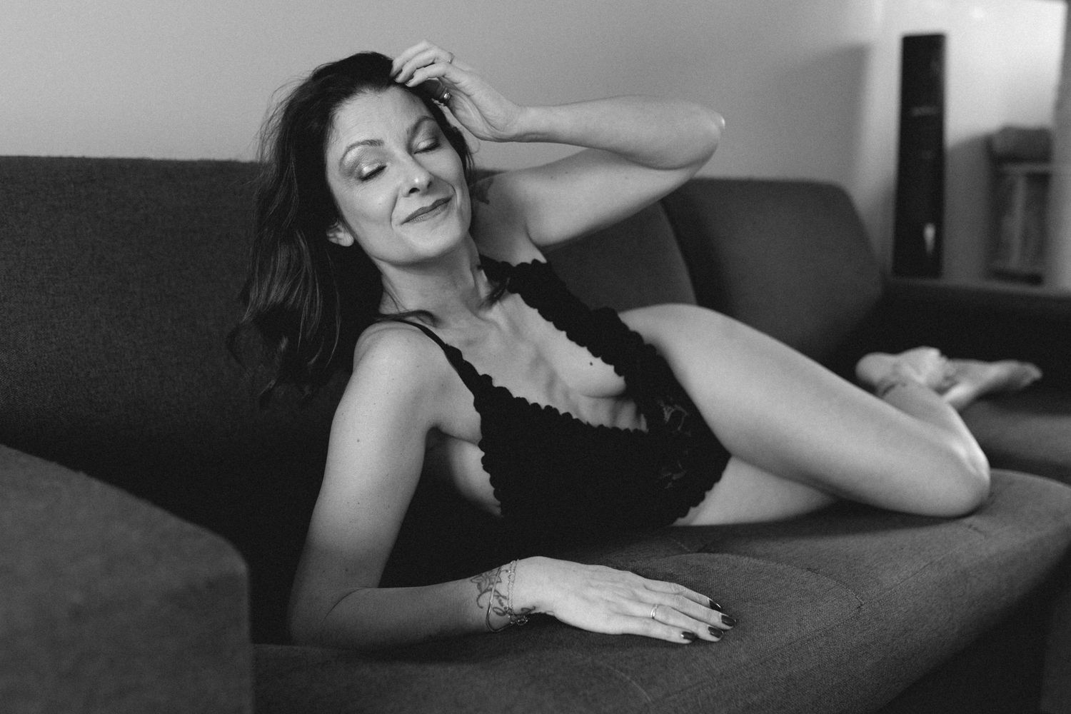 Black and white photo of a woman relaxing on a sofa in a black bodysuit, posing elegantly.
