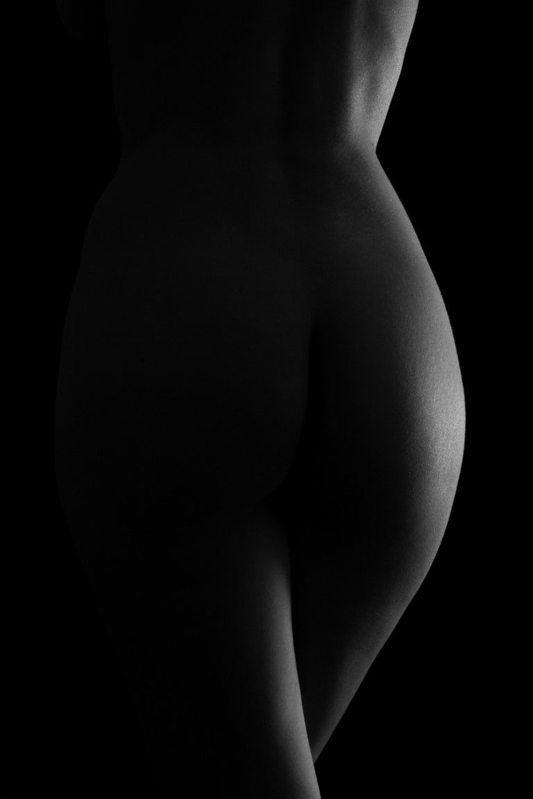Black and white artistic nude photography highlighting feminine curves with dramatic low-key lighting against dark background.