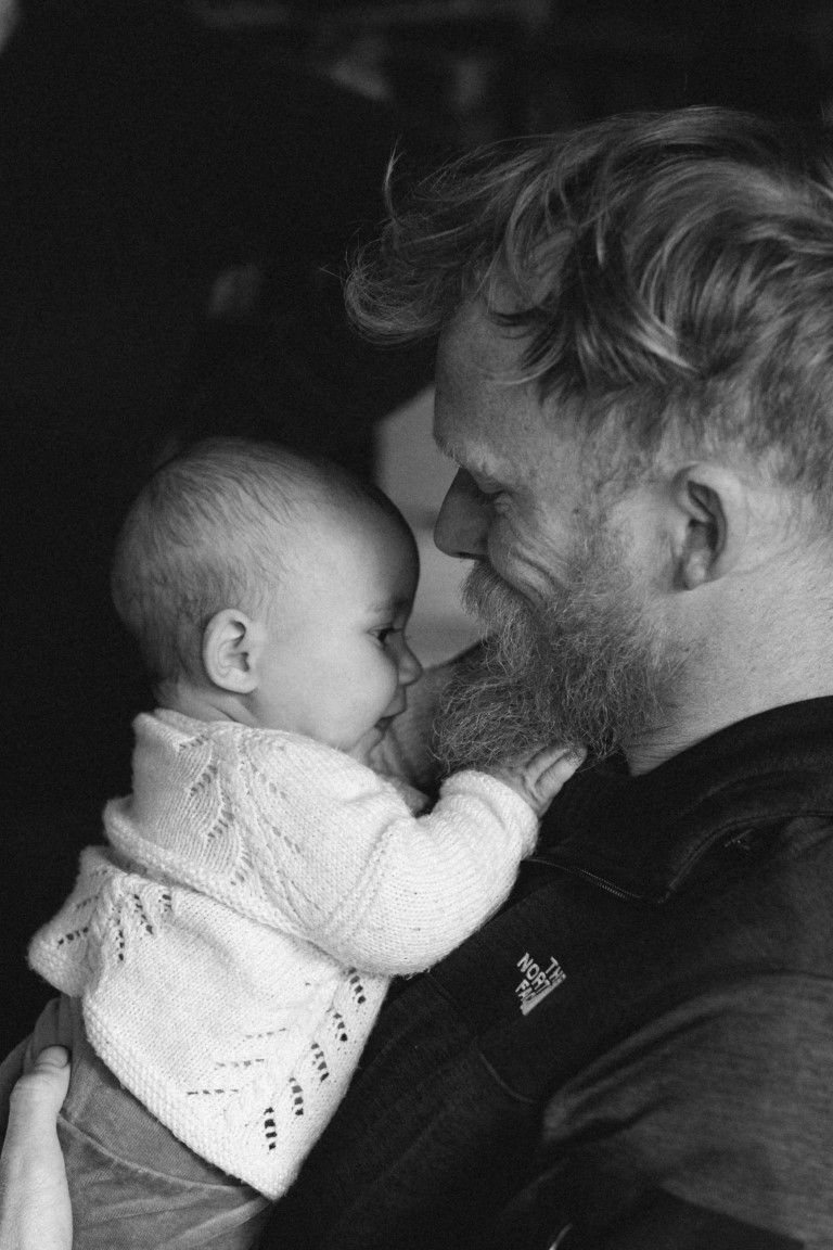 Photo en noir et blanc d'un homme barbu tenant tendrement un petit bébé vêtu d'un pull blanc en tricot et le regardant avec affection.