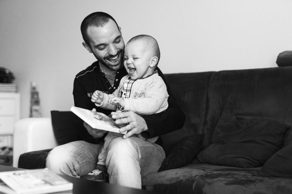 Photo en noir et blanc d'un homme souriant assis sur un canapé tenant un bébé rieur, lisant un livre ensemble.