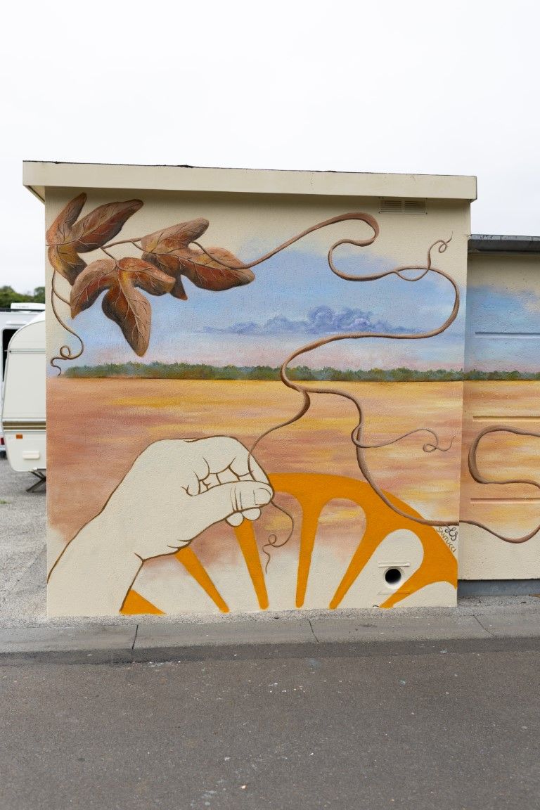 Colorful street art mural on a wall depicting a hand, sun wheel, desert landscape, and seed pods in warm earthy tones.