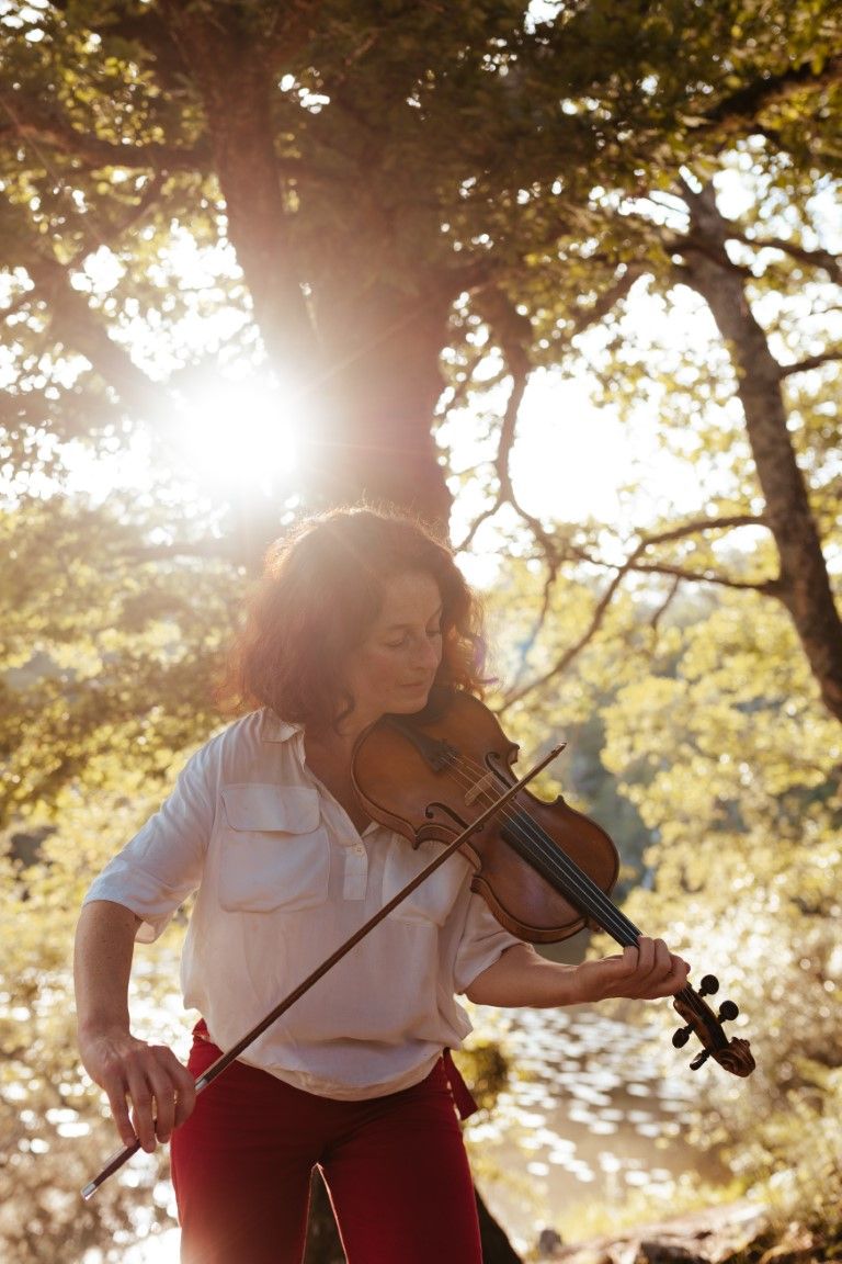 Woman in white shirt playing violin outdoors under sunlit trees with golden light streaming through leaves.