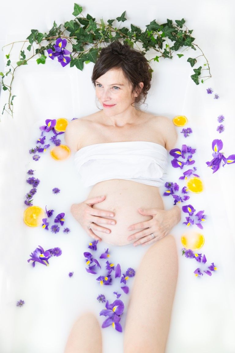 Femme enceinte se détendant dans un bain de lait entourée d'iris violets et de fleurs jaunes.