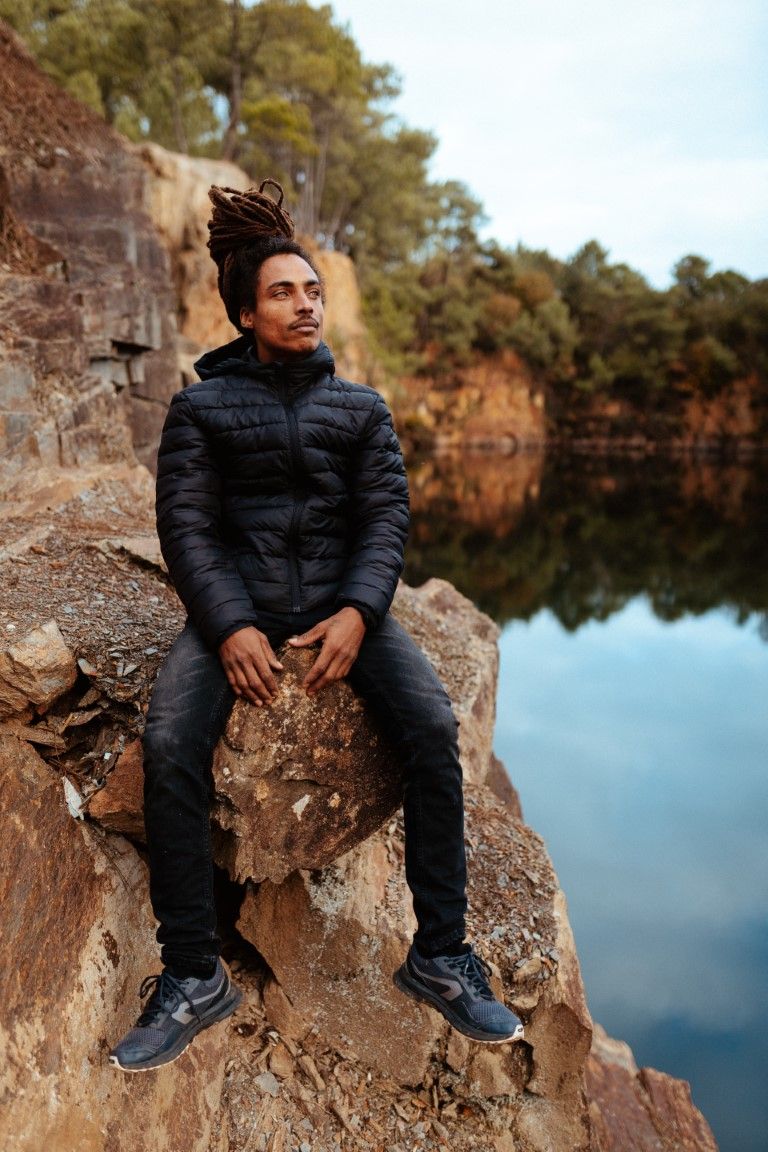 Jeune homme aux dreadlocks assis au bord d'une falaise rocheuse surplombant un lac aux eaux miroitantes.