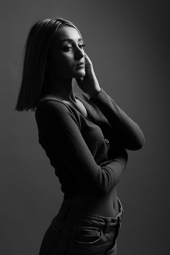 Portrait en noir et blanc, en studio, d'une jeune femme aux cheveux lisses touchant son visage, l'air pensif.