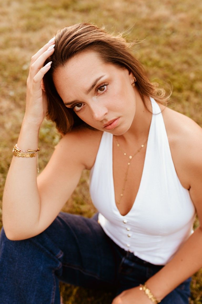 Jeune femme brune en haut blanc dos nu, assise sur l'herbe et portant des bijoux en or.