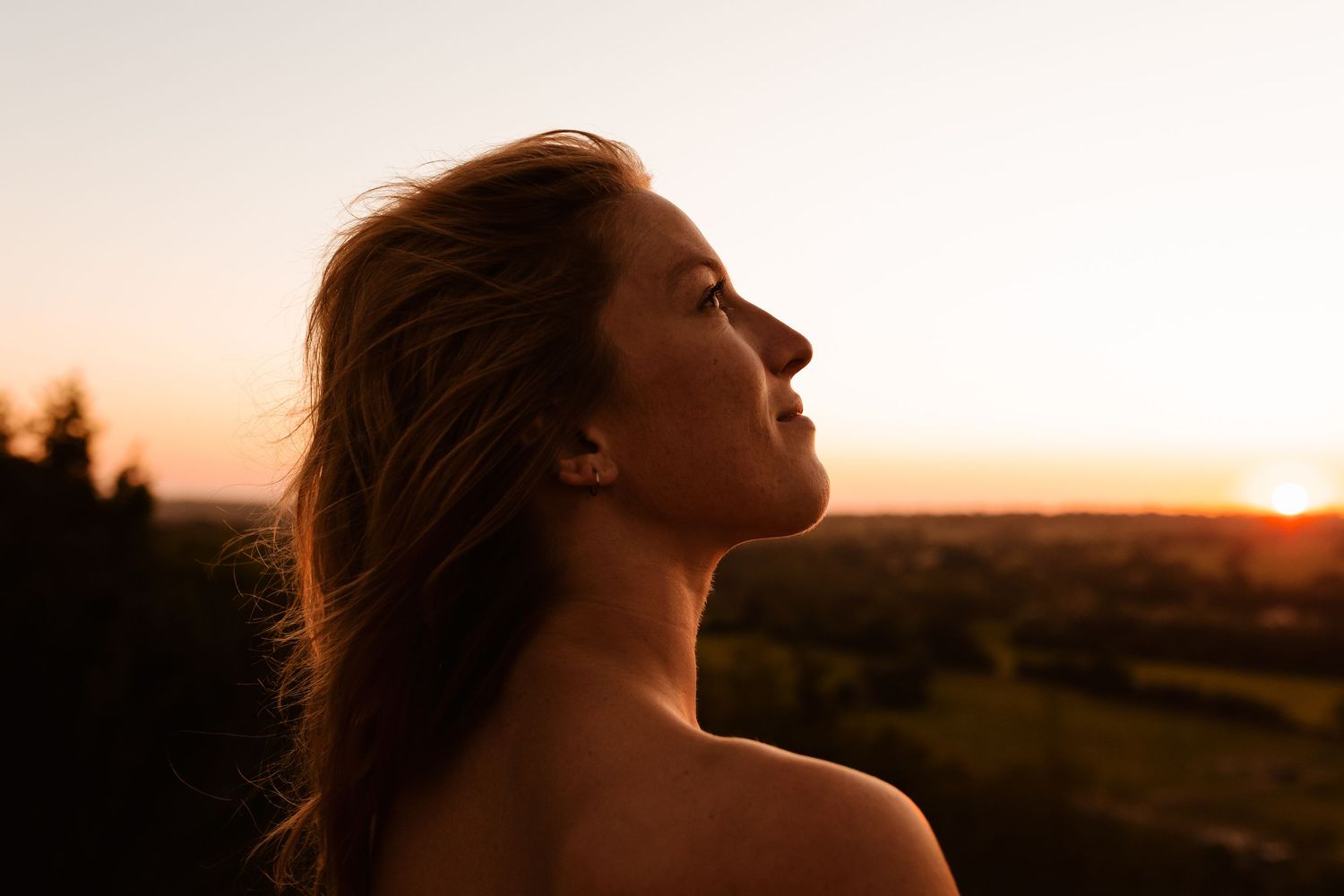 Woman with bare shoulders gazes upward during golden sunset, hair flowing, with scenic landscape in background.
