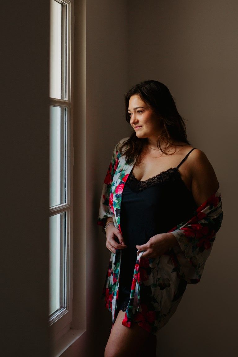 Brunette woman in black lace camisole and floral robe standing by a window in soft light.