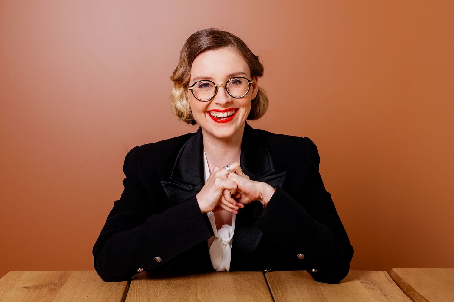 Smiling blonde woman with red lips and glasses sitting at wooden table in black blazer.