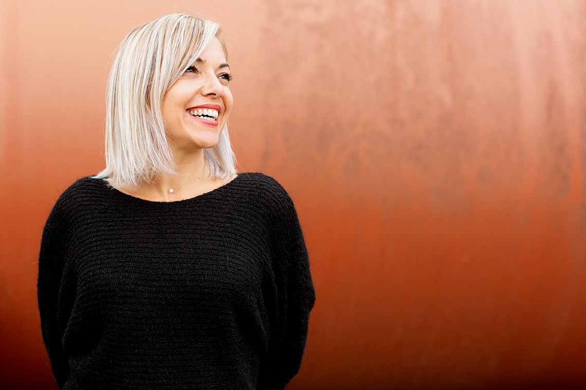 Platinum blonde woman in black sweater laughing against warm terracotta-colored wall background.