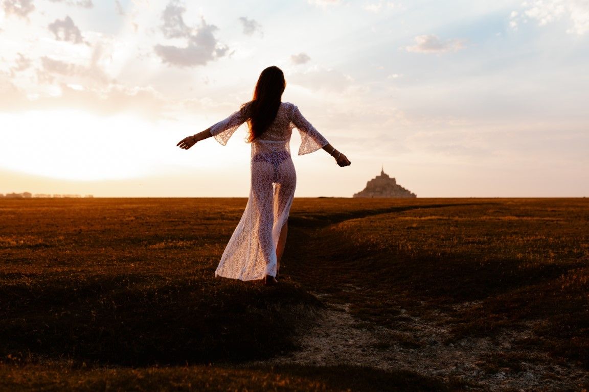 Woman in flowing white dress standing with arms outstretched at golden sunset near Mont Saint-Michel.