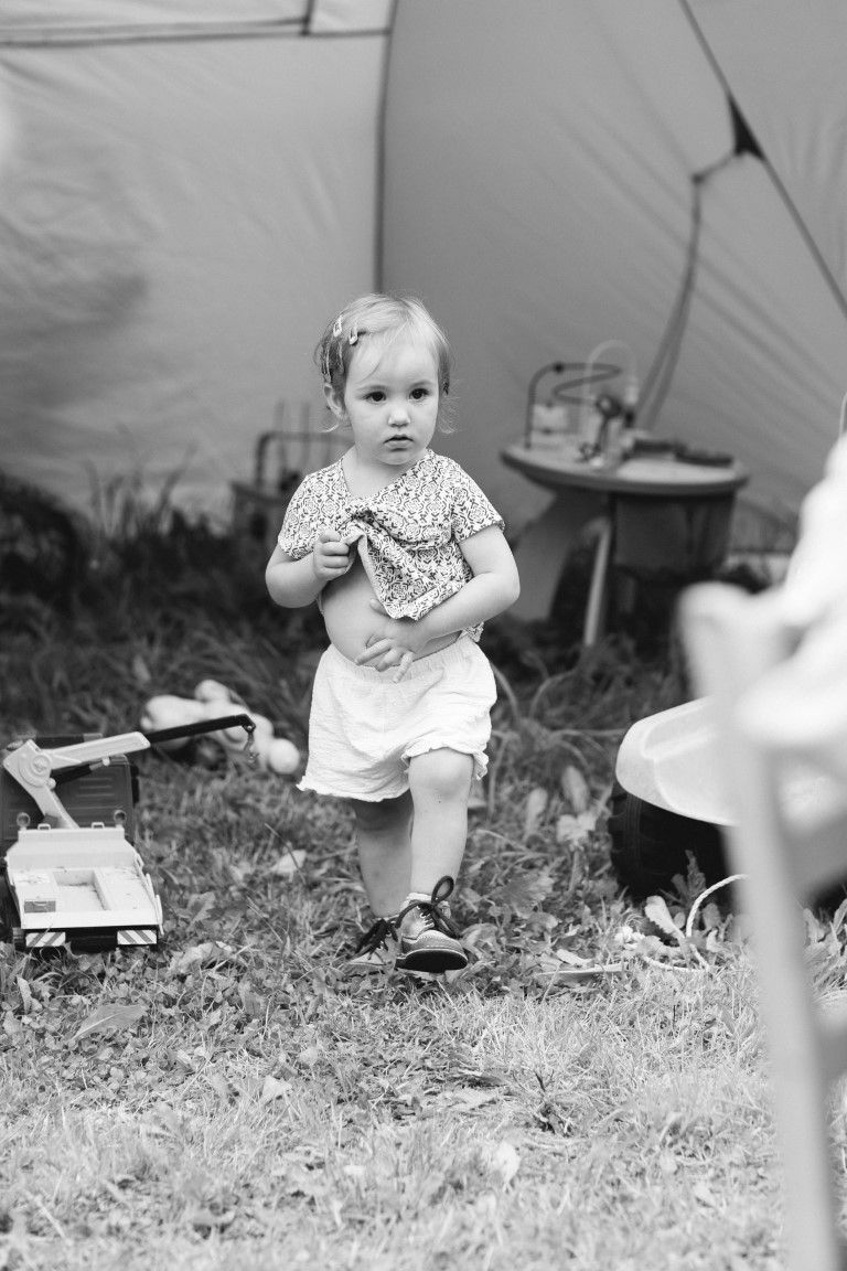 Photo en noir et blanc d'une petite fille courant à l'extérieur près d'une tente entourée de jouets sur l'herbe.