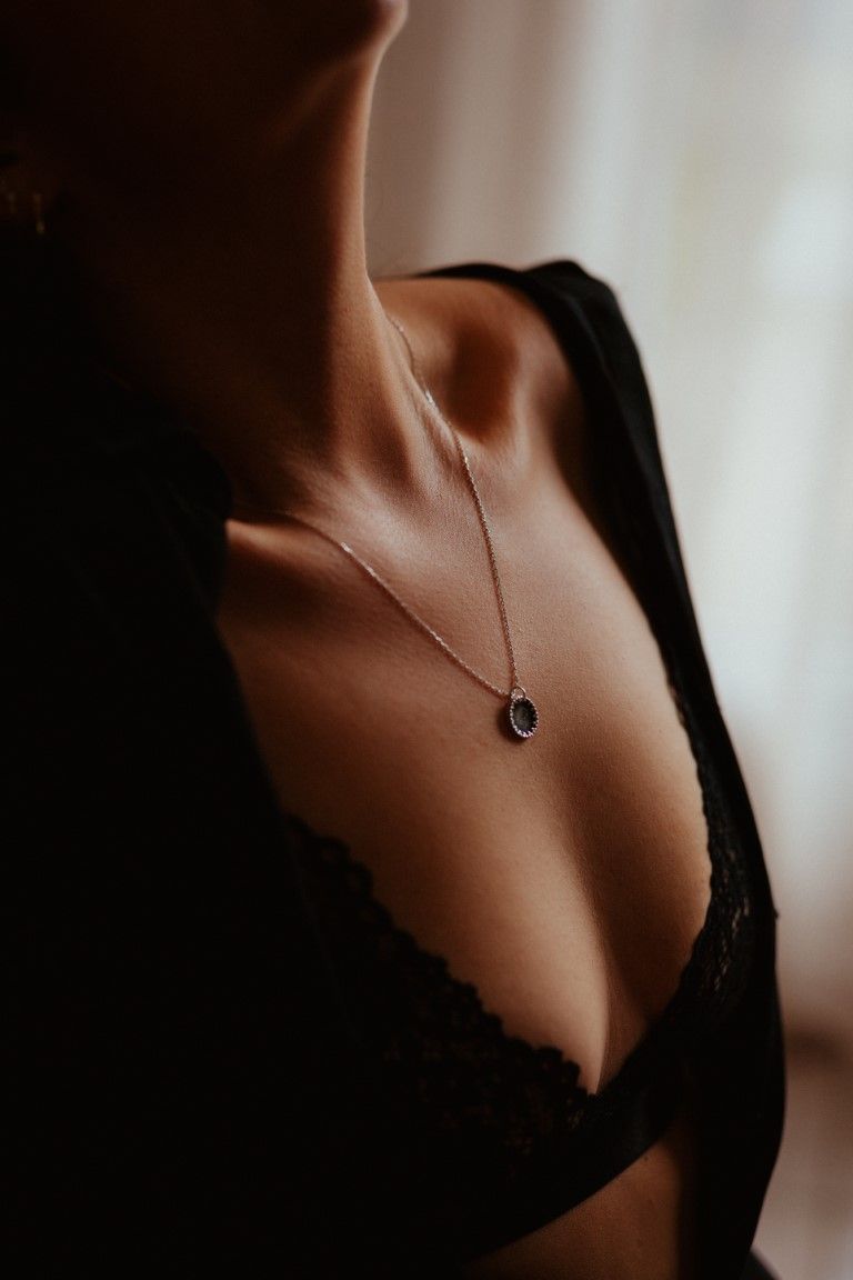 Close-up of a woman wearing black lace lingerie and a delicate oval pendant necklace in soft lighting.