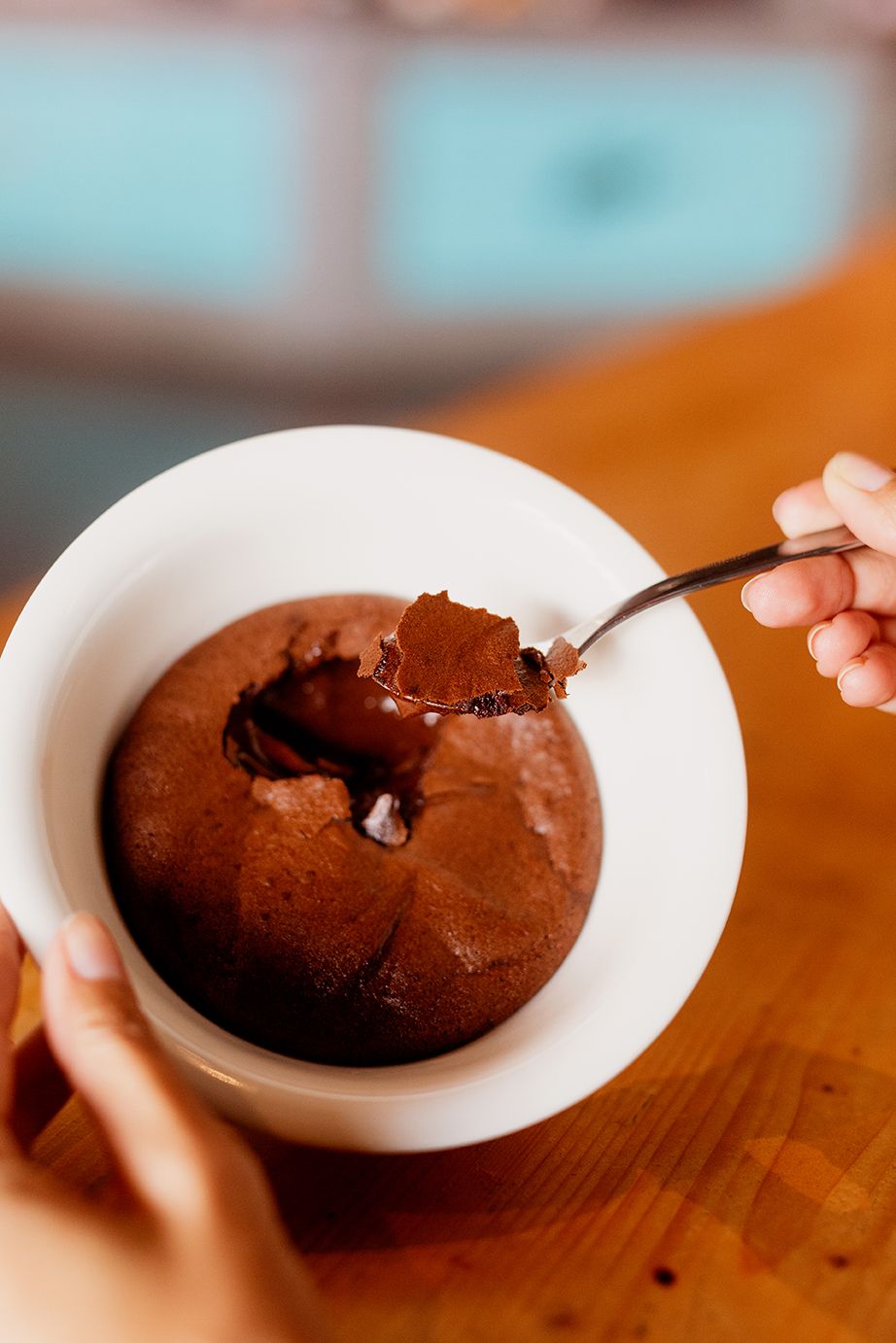 A hand holds a white bowl with a chocolate lava cake, spoon lifting a piece revealing molten center.