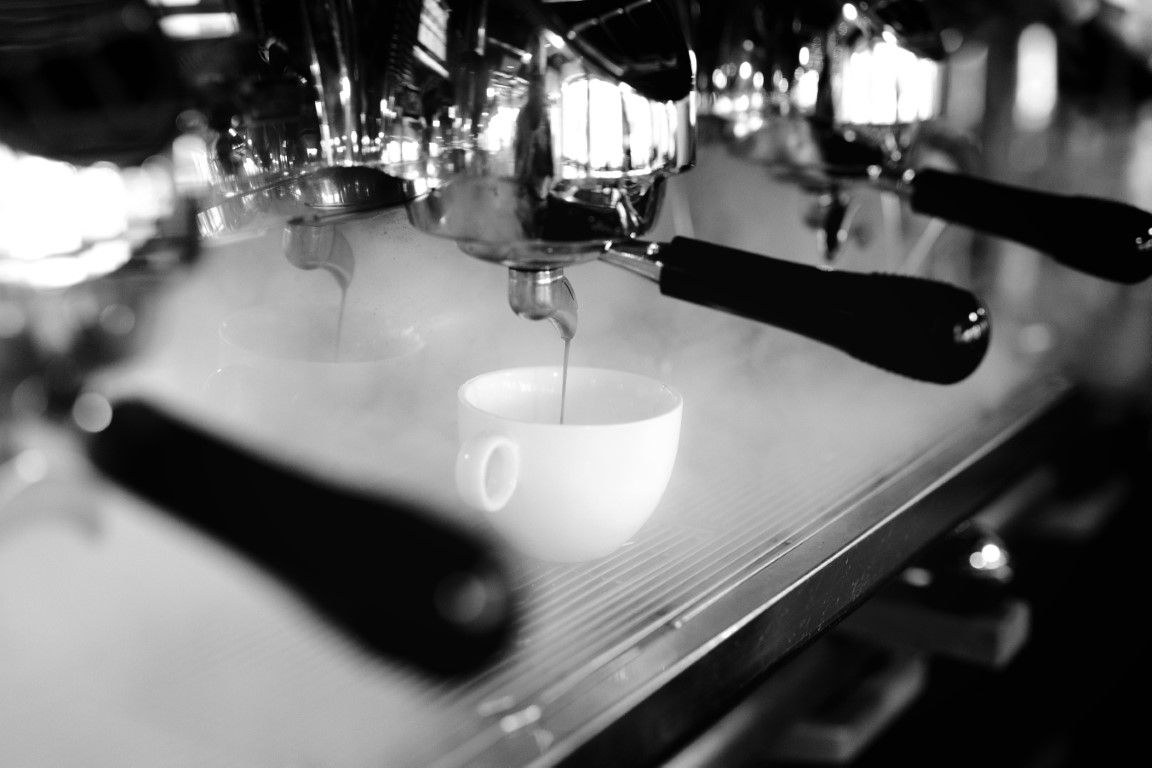 Black and white photo of espresso machine brewing coffee into a white cup with steam rising.