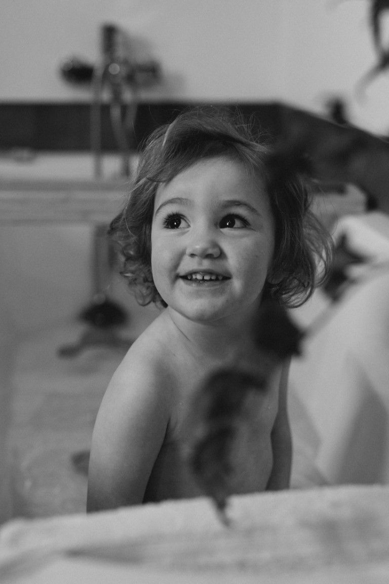 Enfant souriant aux cheveux bouclés assise dans une baignoire, photo en noir et blanc.