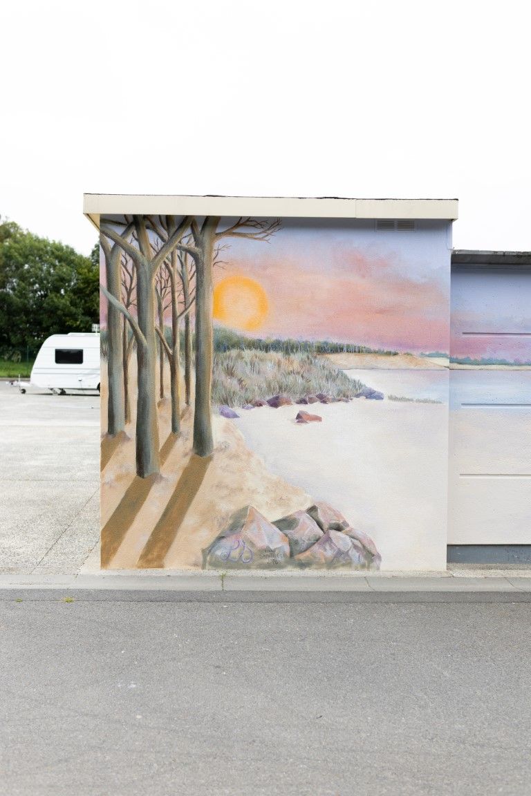 A colorful mural of a coastal sunset with bare trees and rocky shoreline painted on a building wall.